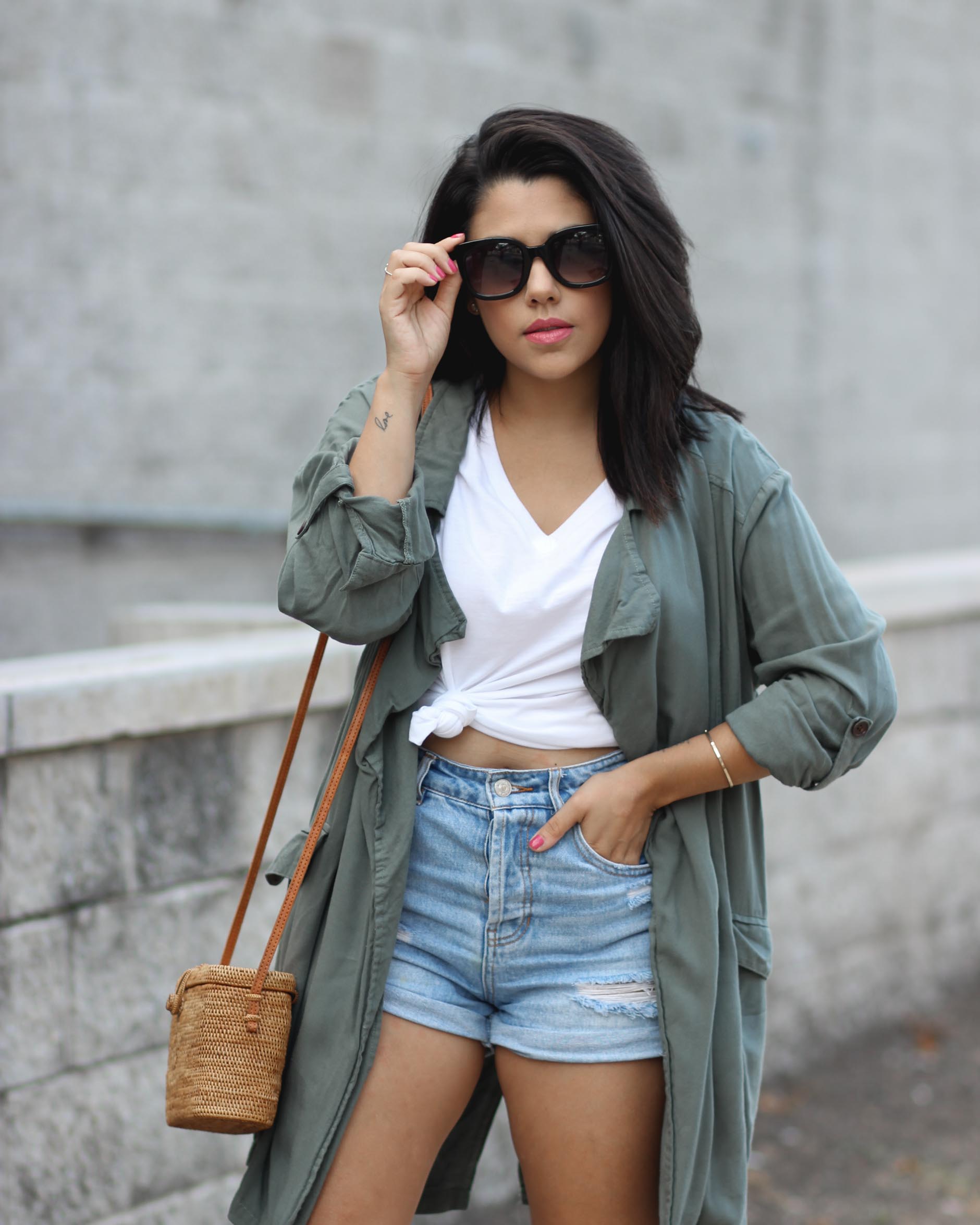 lifestyle blogger naty michele wearing a knotted tee with a trench coat and denim shorts holding her sunglasses